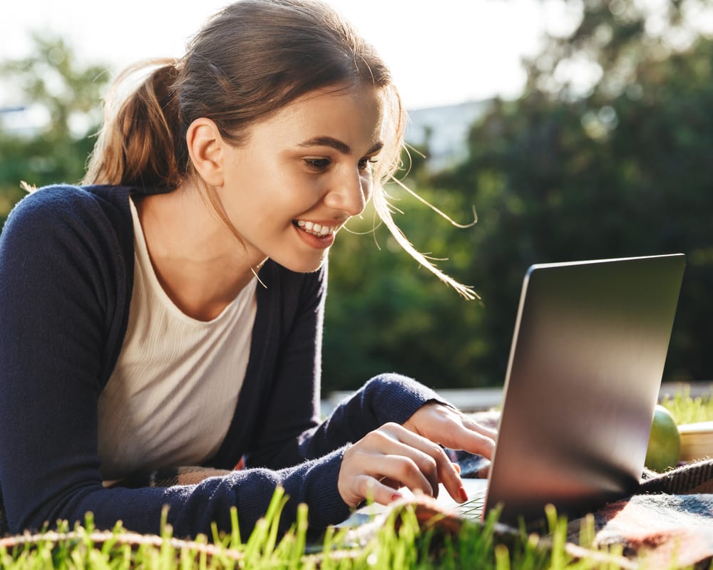 Young happy woman lying on a blanket in a park and typing on a laptop