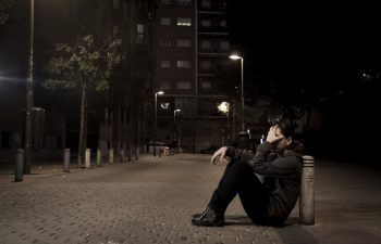 A depressed person sitting on the pavement and covering their face with a hand.