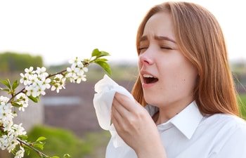 A sneezing young woman standing next to a flowering tree.